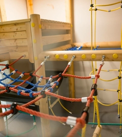 Ein Holzbett und ein Kletternetz prägen den Kinder-Spielbereich mit bunten Seilen.