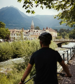 A cyclist rides along a riverside path, with Naturns, a bridge and mountains in the background.