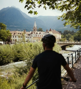 A cyclist rides along a riverside path, with Naturns, a bridge and mountains in the background.