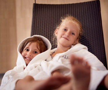Zwei Kinder sitzen eingekuschelt in Bademänteln auf einer Liege im Family Spa.