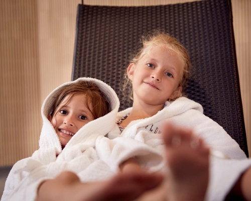 Zwei Kinder sitzen eingekuschelt in Bademänteln auf einer Liege im Family Spa.