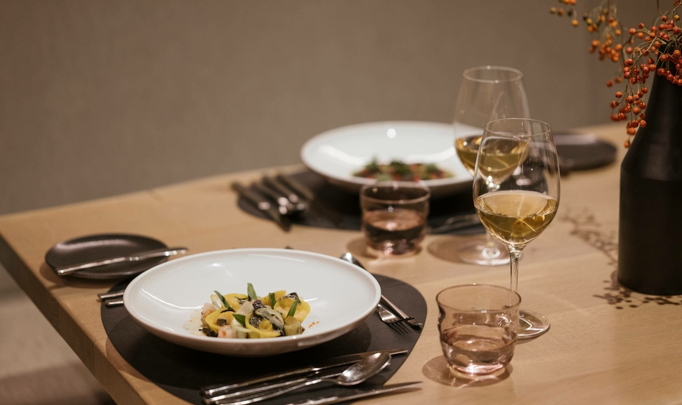 Ein gedeckter Tisch im Hotelrestaurant mit weißem Geschirr, Weingläsern und dunklen Platzsets.