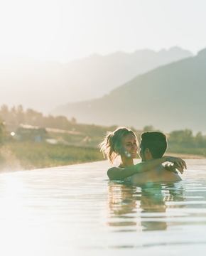Ein Paar steht im Infinity-Pool mit Blick auf die Berge im warmen Licht der Abendsonne.