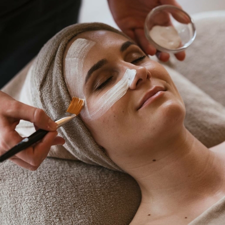 A woman receives a facial treatment in the spa while cream is applied with a brush.