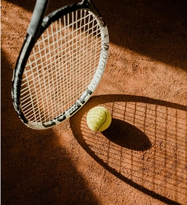 A tennis ball lies beside a racket on a red clay court surface.