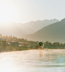 Eine Person steht im Infinity-Pool mit Blick auf die Berge im warmen Abendlicht.