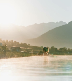 Eine Person steht im Infinity-Pool mit Blick auf die Berge im warmen Abendlicht.