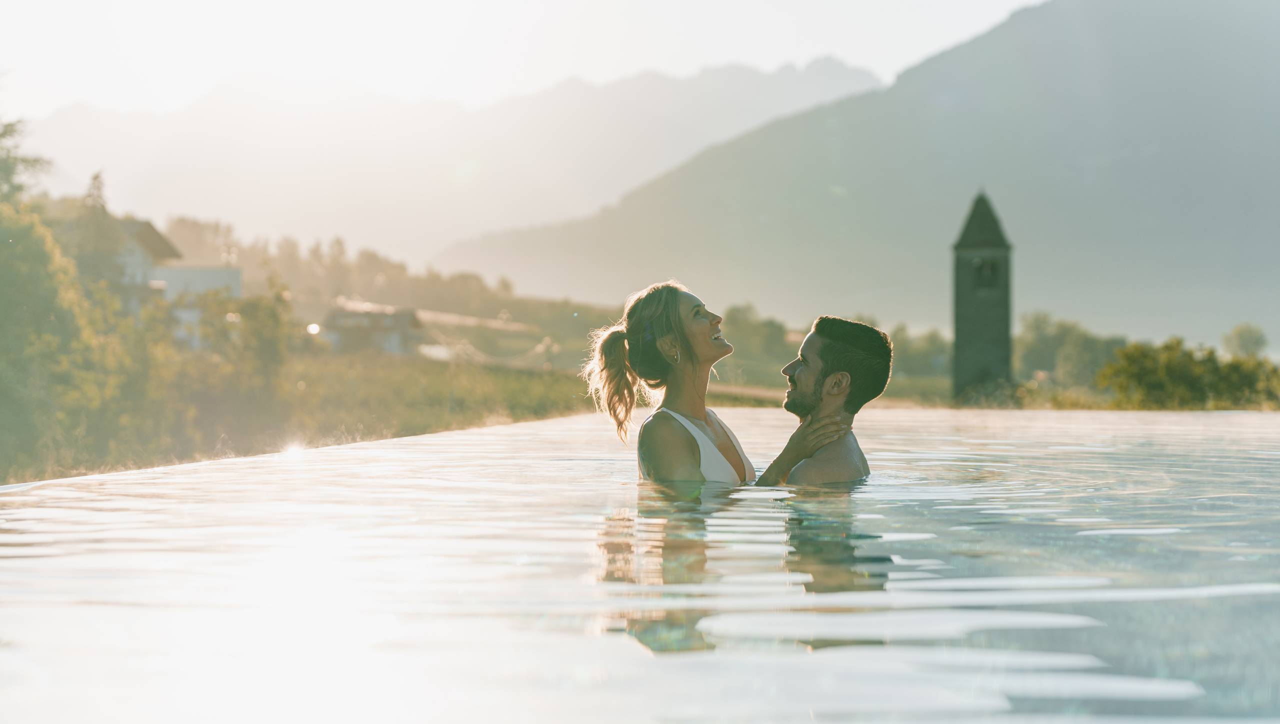 A couple stands in an outdoor pool with mountains and a church tower in the background.
