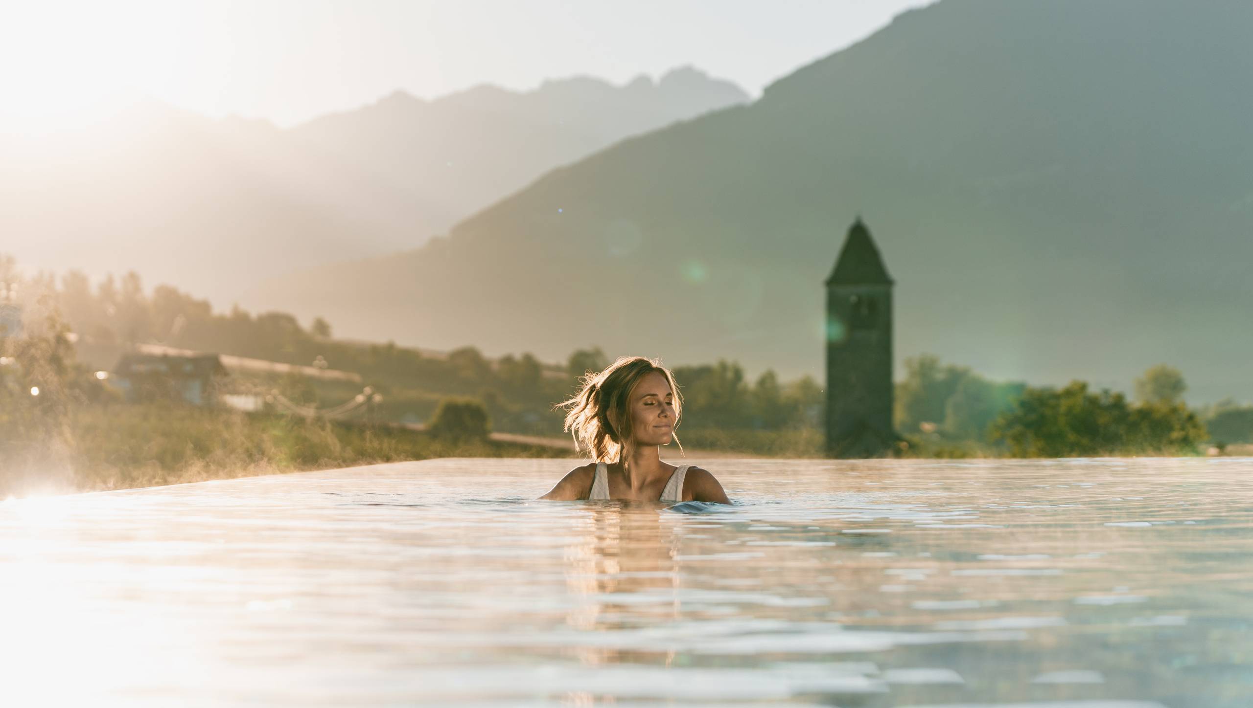 Eine Frau steht im Infinity-Pool vor einer Bergkulisse im warmen Abendlicht.